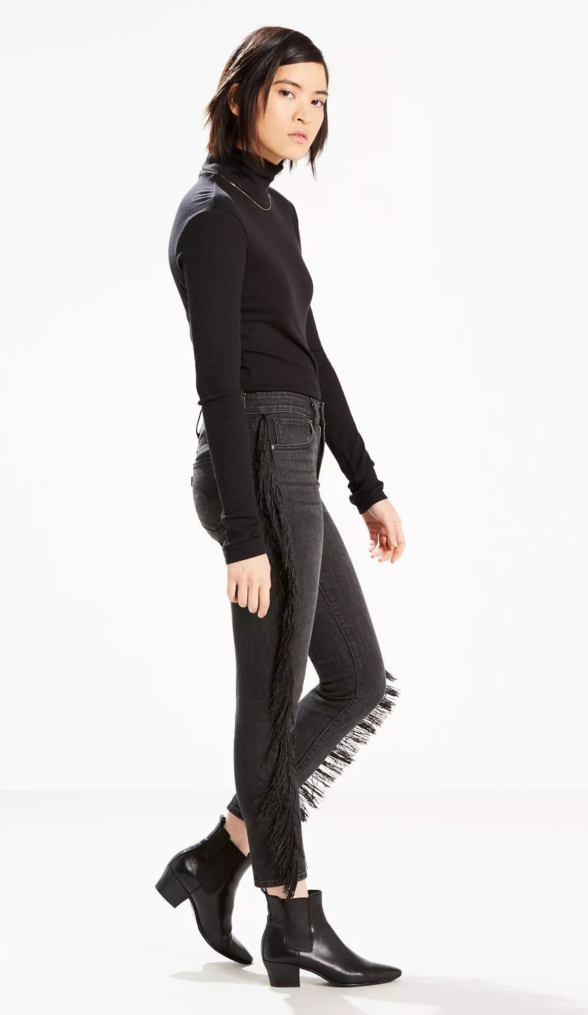 A woman models a fitted black turtleneck and Levi's Limited 721 Fringe-Trim Skinny Ankle Jeans Black Size 24, showcasing a contemporary casual style against a white background.