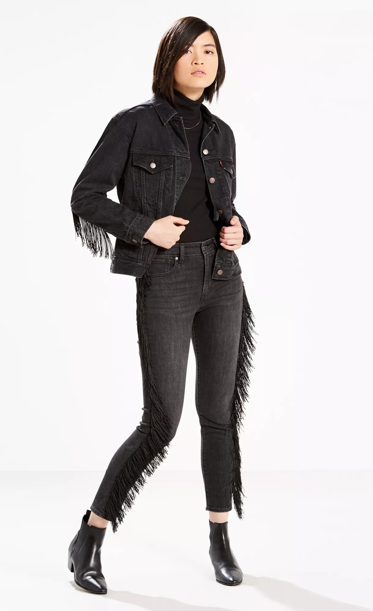 A stylish woman poses confidently, wearing a fringe-detailed black Levi's Limited 721 Fringe-Trim Skinny Ankle Jeans Black Size 24 denim jacket and matching high rise skinny fringe trim jeans, paired with a simple top and ankle boots, embodying a modern.